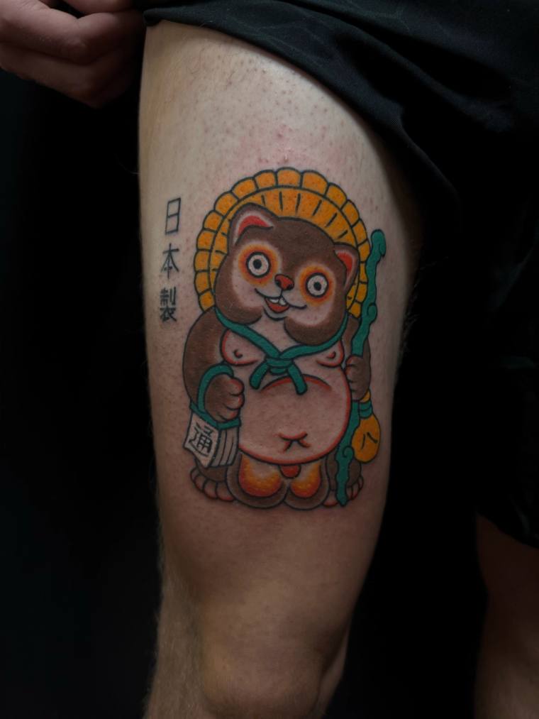 Color irezumi tattoo on thigh depicting a smiling tanuki holding a sake bottle marked with the kanji for fortune, framed by a