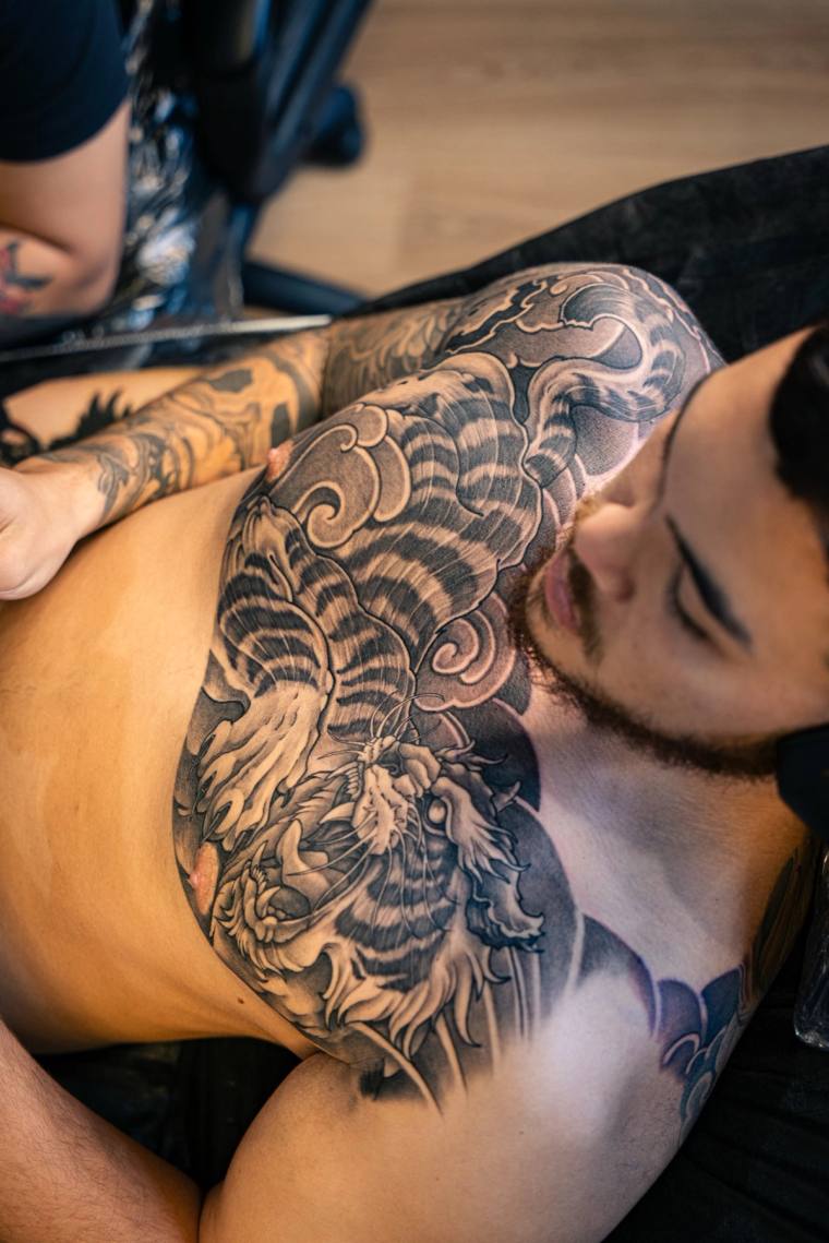 A large black and grey Japanese irezumi tattoo of a tiger and dragon head with clouds and waves across a man's chest and shou