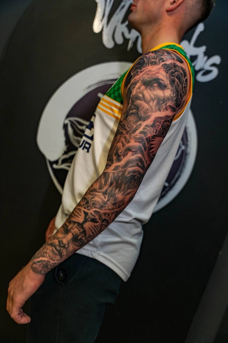 Black and grey realism full sleeve tattoo featuring a classical bearded god at the shoulder, a roaring lion mid-arm, and a wo