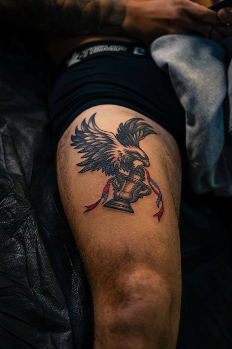 Neo-traditional tattoo on a thigh featuring a black eagle with spread wings perched atop a chess rook, with red and blue ribb