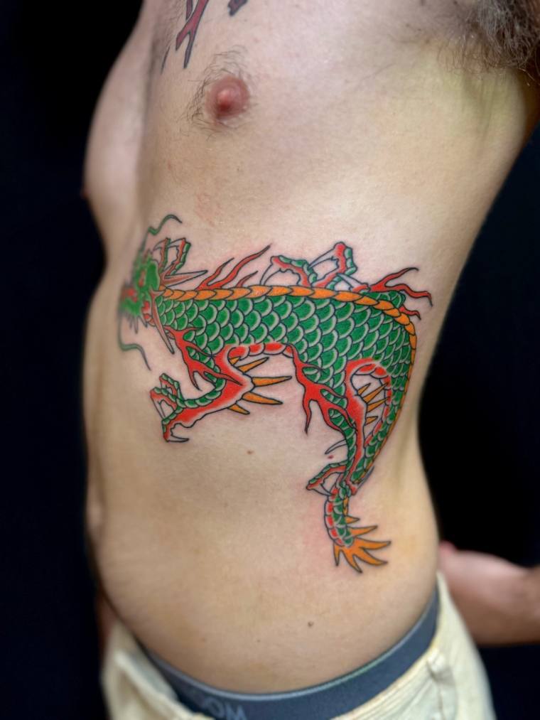 Colourful green-scaled Asian dragon with orange flames inked on a man's ribs in traditional irezumi style.