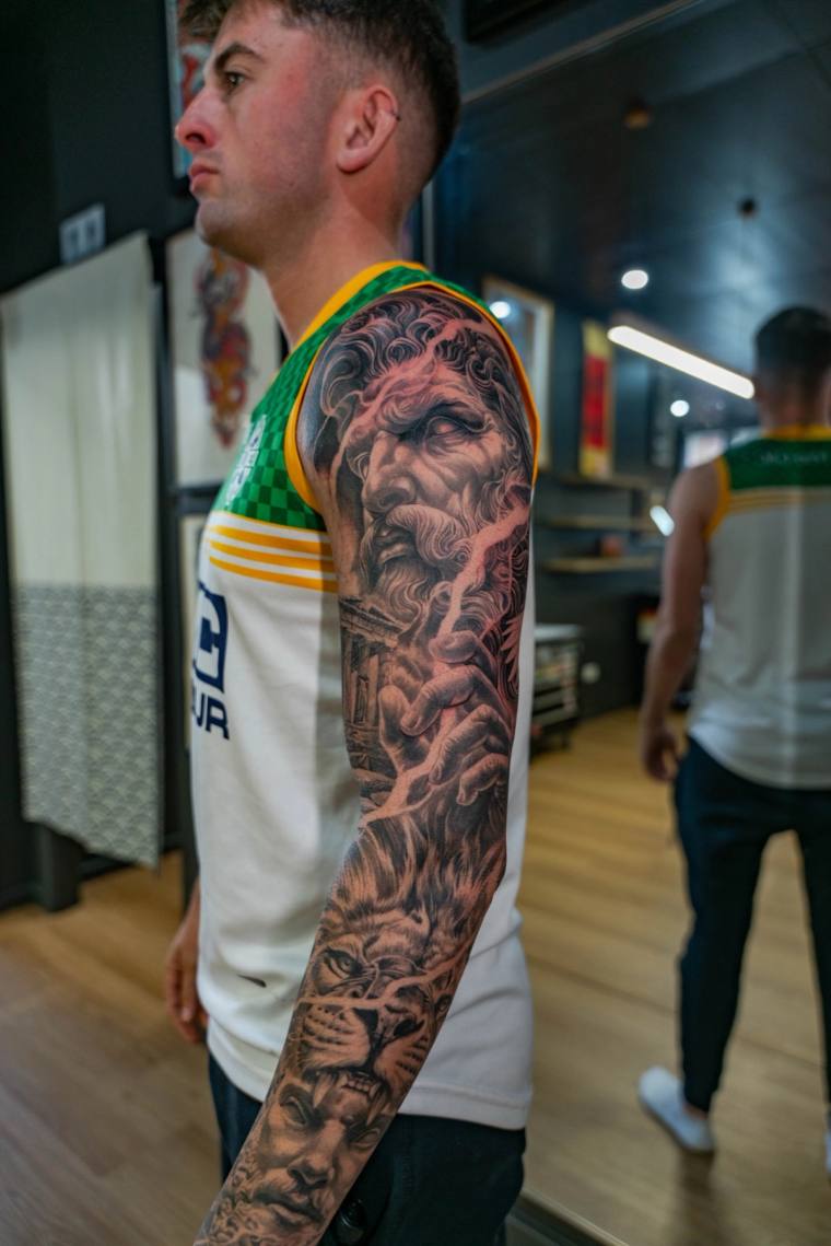 Profile view of a man displaying a finished black and grey full sleeve tattoo featuring a classical bearded deity figure on t