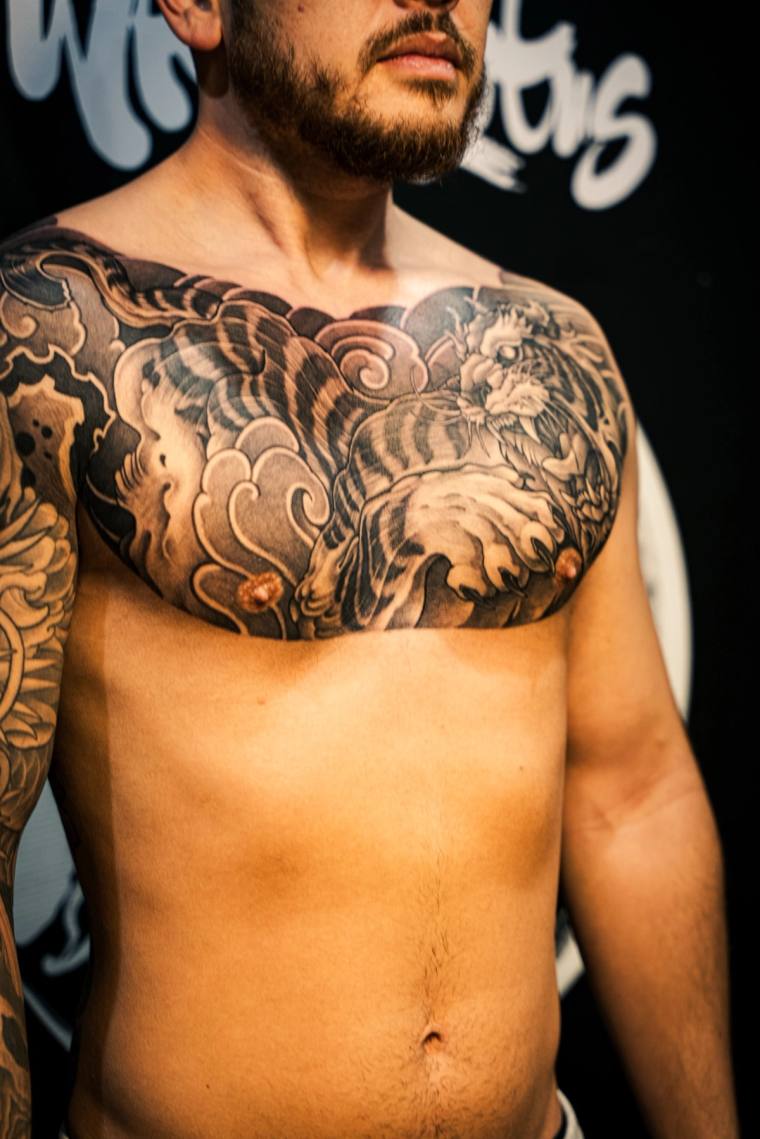 Bearded man displaying a finished black and grey irezumi chest piece featuring a tiger, wind clouds, and crashing waves acros