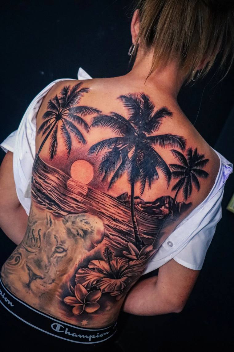 Large black and grey back tattoo featuring palm trees against a setting sun, a lion portrait, hibiscus and plumeria flowers, 