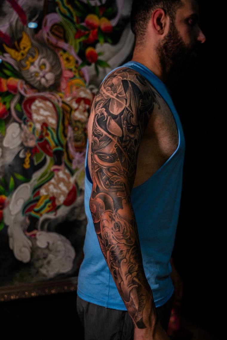 Profile view of a man in a blue tank top showing off a finished black and grey full sleeve tattoo featuring a samurai mask, k