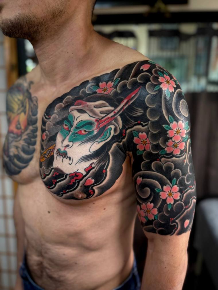 Finished irezumi half sleeve featuring a green hannya mask with red accents, framed by black kumo clouds and pink cherry blos