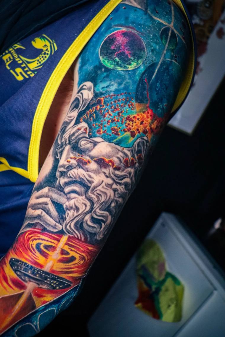 Vibrant cosmic sleeve tattoo featuring a classical deity face, a planet containing a figure, a UFO beaming light, and swirlin
