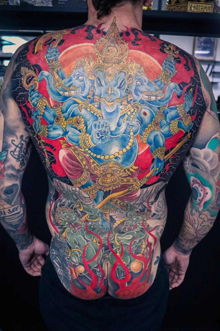 Vibrant full-back irezumi tattoo of blue multi-armed Ganesha against a red sun, with green dragon and red flames wrapping the