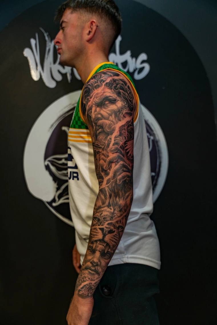 Side profile of a man displaying a finished black and grey full sleeve tattoo featuring a bearded god, lion, and classical po