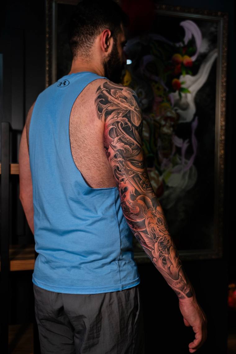 Bearded man in a blue tank top displaying a finished black and grey Japanese irezumi sleeve featuring koi, waves, and clouds 