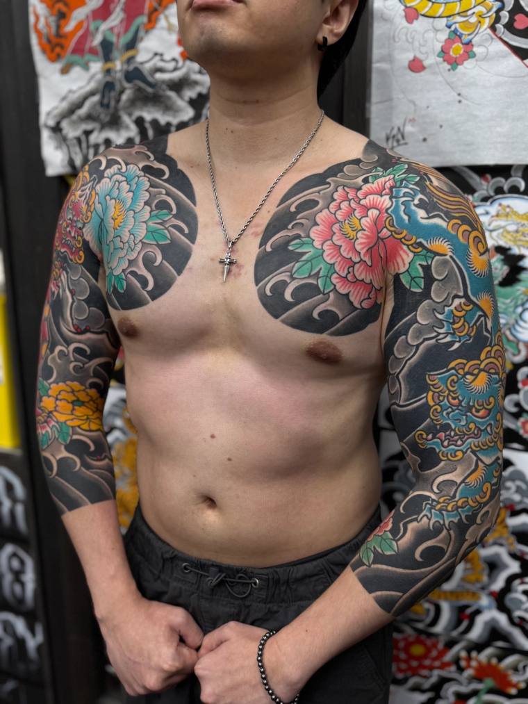 Shirtless man displaying vibrant irezumi full sleeves and chest panels featuring pink peonies, grey wind bars, and a dragon m