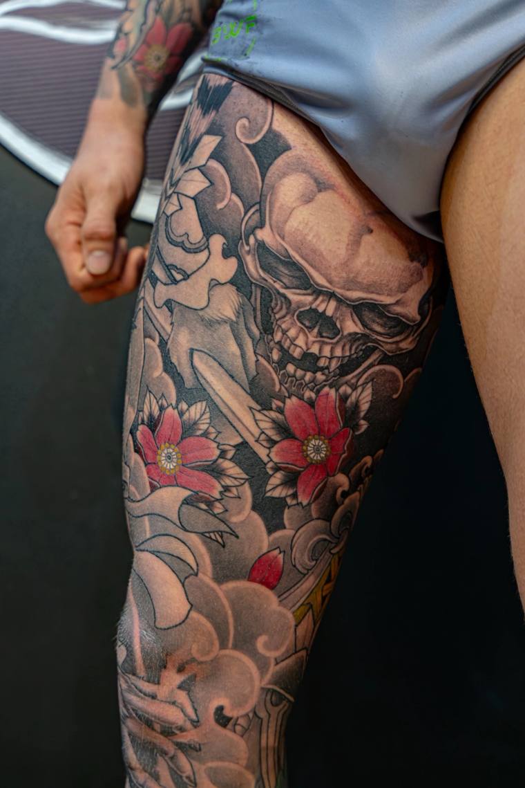 Large greywash skull tattoo surrounded by red cherry blossoms and swirling Japanese-style clouds on a thigh.