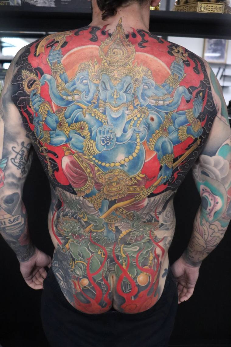 Large irezumi back piece featuring blue multi-armed Ganesha with golden crown and jewelry, red cloud backdrop, green dragon, 