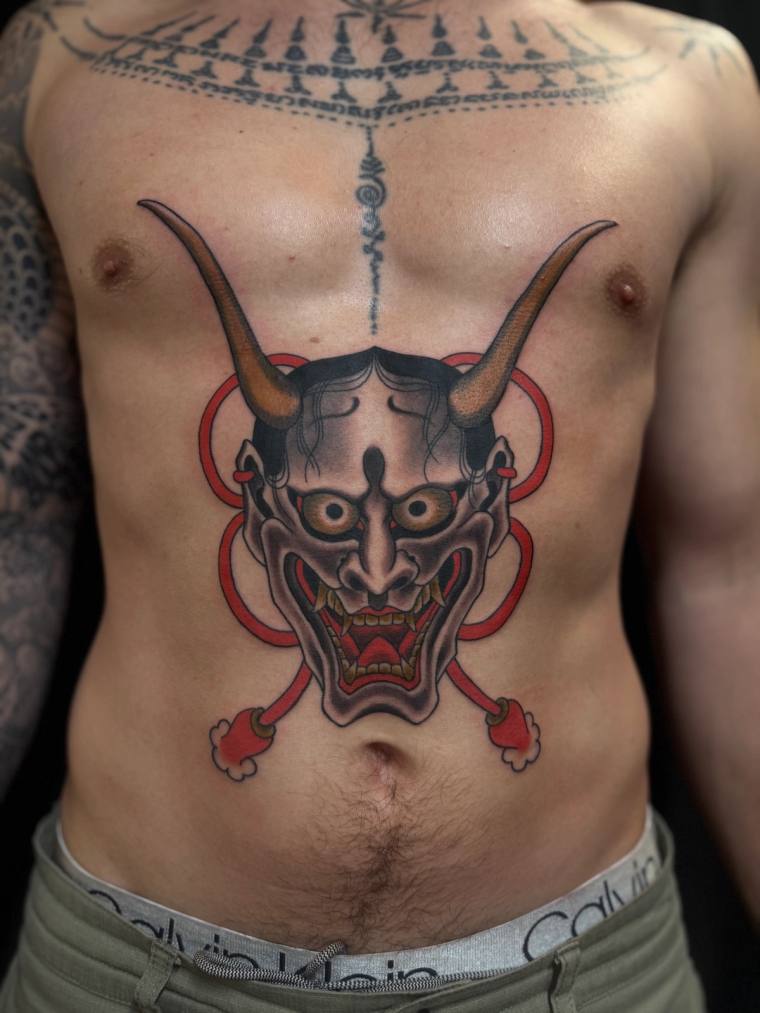 Large colour irezumi hannya demon mask tattoo on a man's abdomen, framed by a red ring, with Sak Yant script across the upper