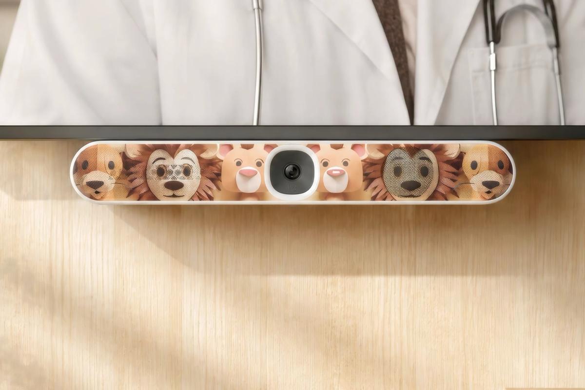 Video bar in a Heckler easy-clean cover printed with friendly animal faces, mounted below a display showing a doctor in a pediatric telehealth room.