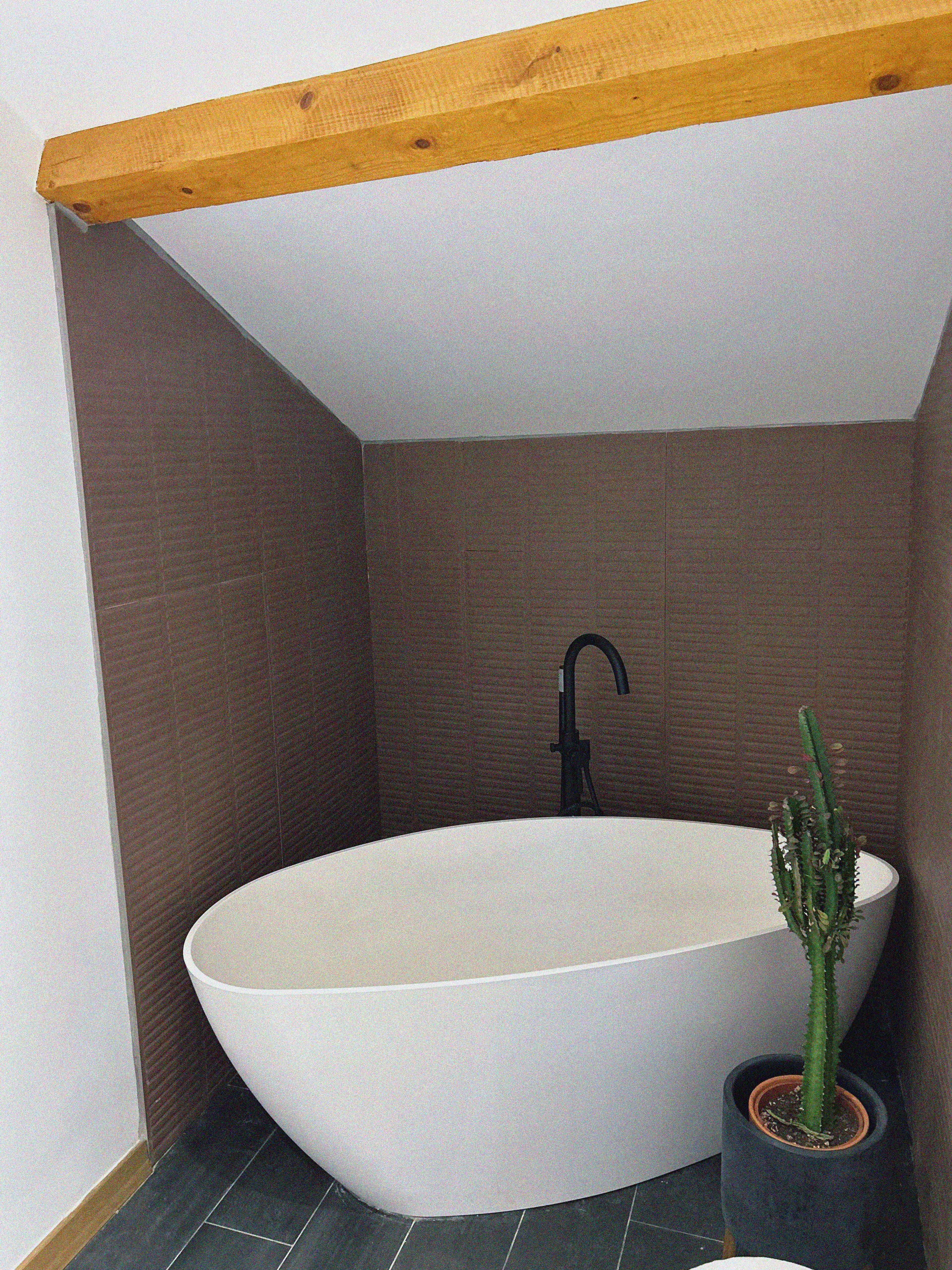 Baignoire îlot dans sa niche de carrelage texturé couleur terre, sous les poutres apparentes