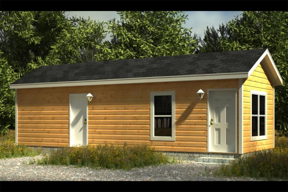 Exterior view of a small manufactured home with wood-look siding, a gable roof, white trim, and a covered front porch with railing. Surrounded by trees and gravel landscaping.