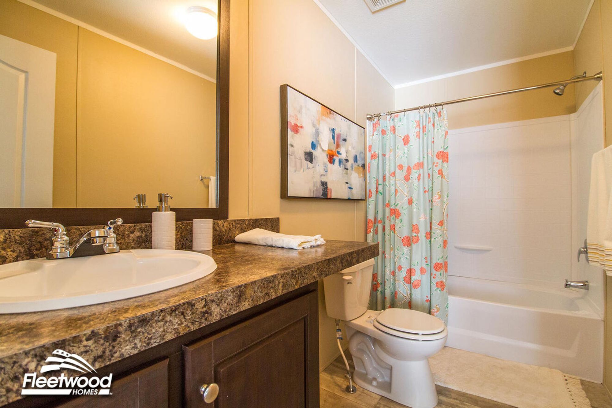 Bathroom with a brown marble-style countertop, single sink, large mirror, white toilet, and bathtub-shower combo with a floral shower curtain. Light tan walls and decorative wall art complete the space.