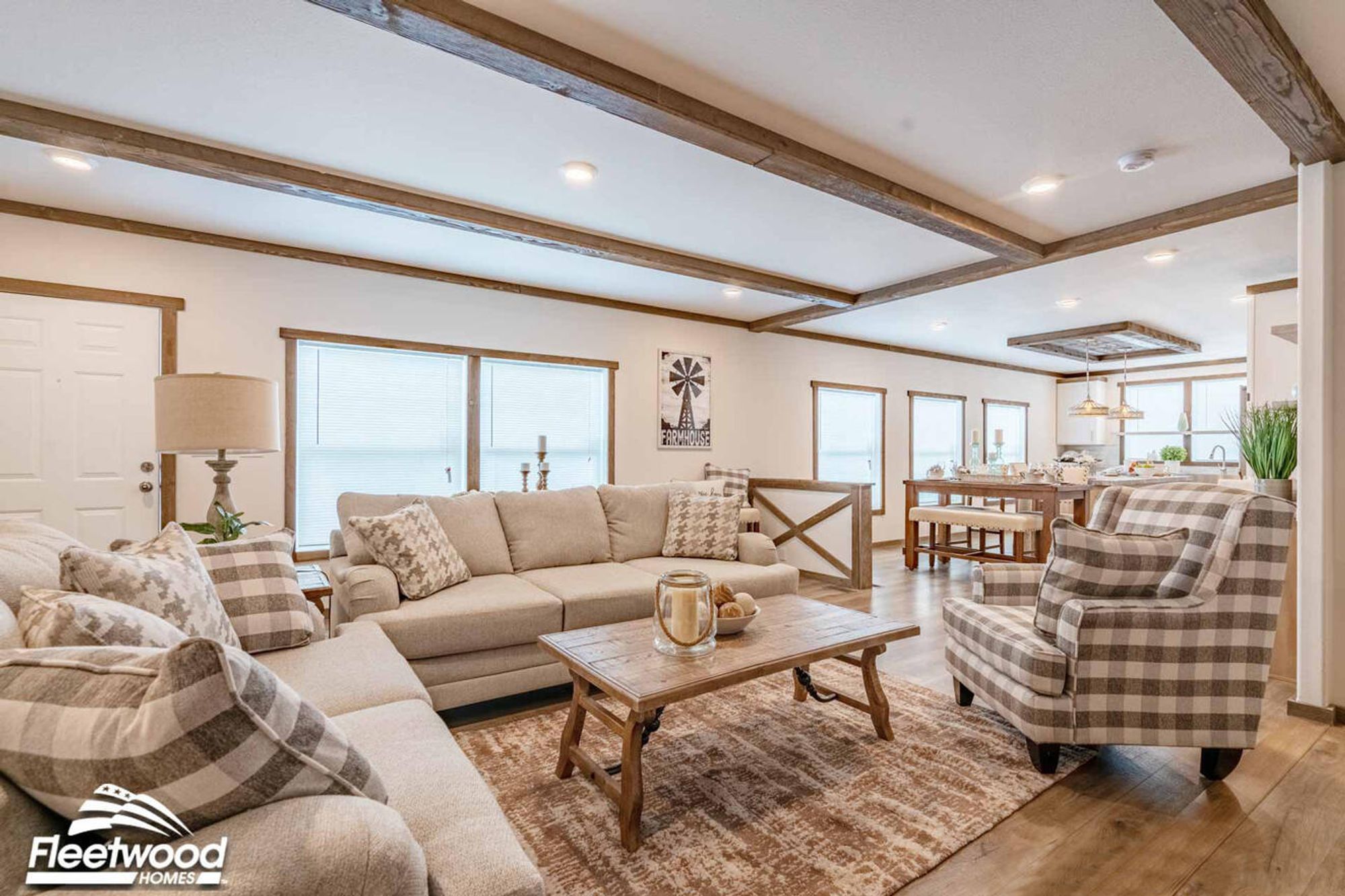 Cozy living room with wood-beam ceiling accents, neutral-toned sofas with patterned pillows, a plaid armchair, a rustic coffee table, and a wood-paneled fireplace wall with a mounted TV.
