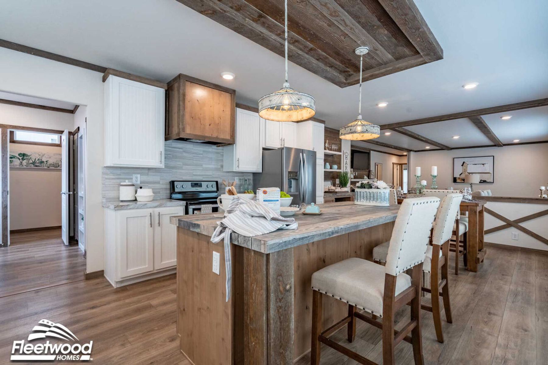 Open-concept kitchen and dining area with a rustic wood island, pendant lighting, wood-look flooring, and large windows, flowing into a living room with a sectional sofa.
