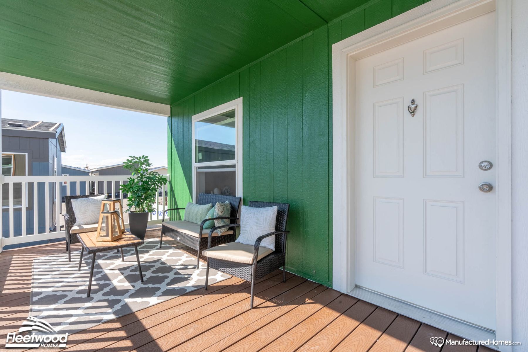 Covered front porch with green siding, outdoor seating, a small table, rug, and potted plant. White front door and railing, with neighboring homes visible in the background.