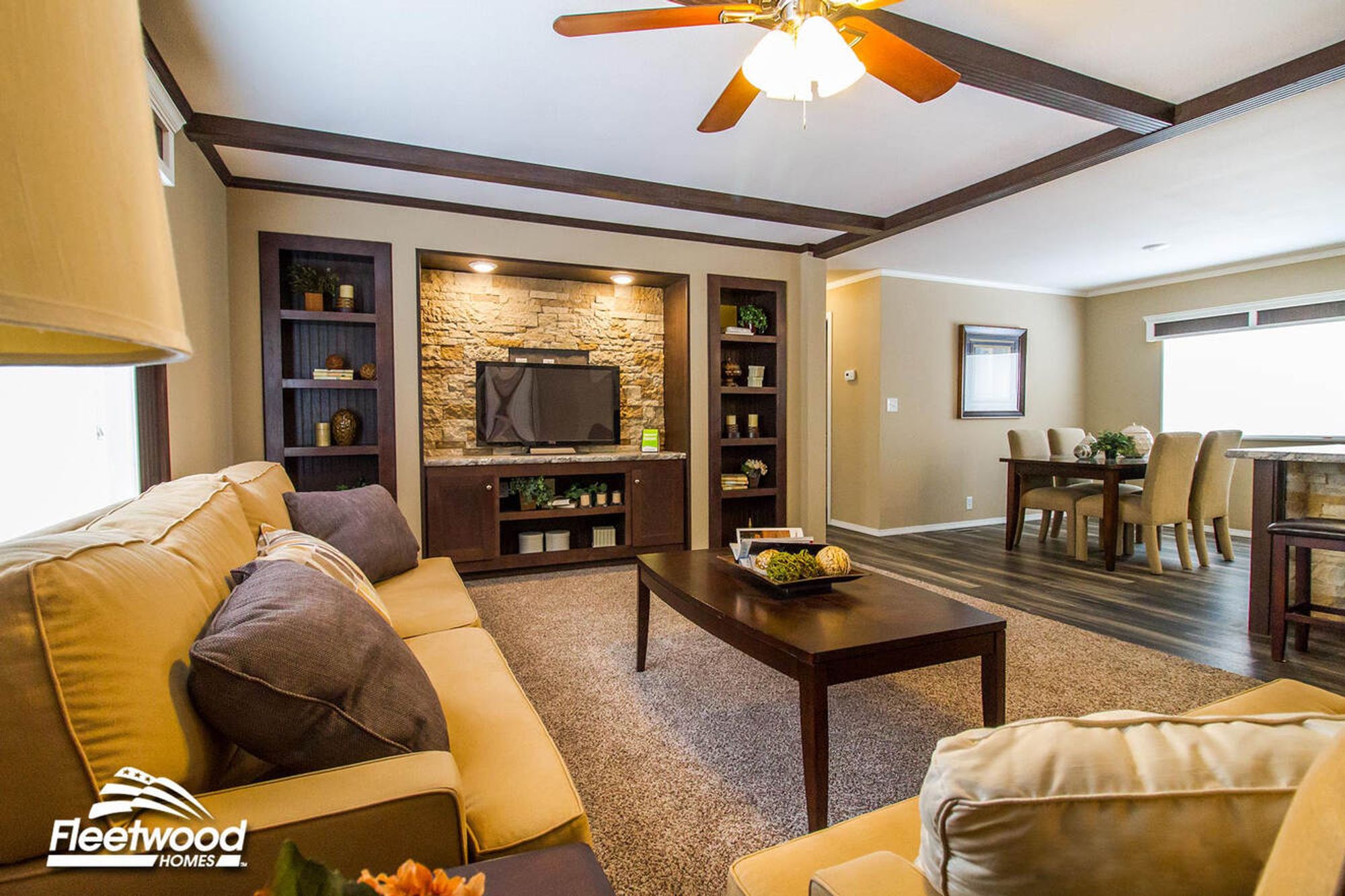Open living and dining area with tan sofas, wood coffee table, ceiling fan, and a stone-accented entertainment center across from a dining table with upholstered chairs.