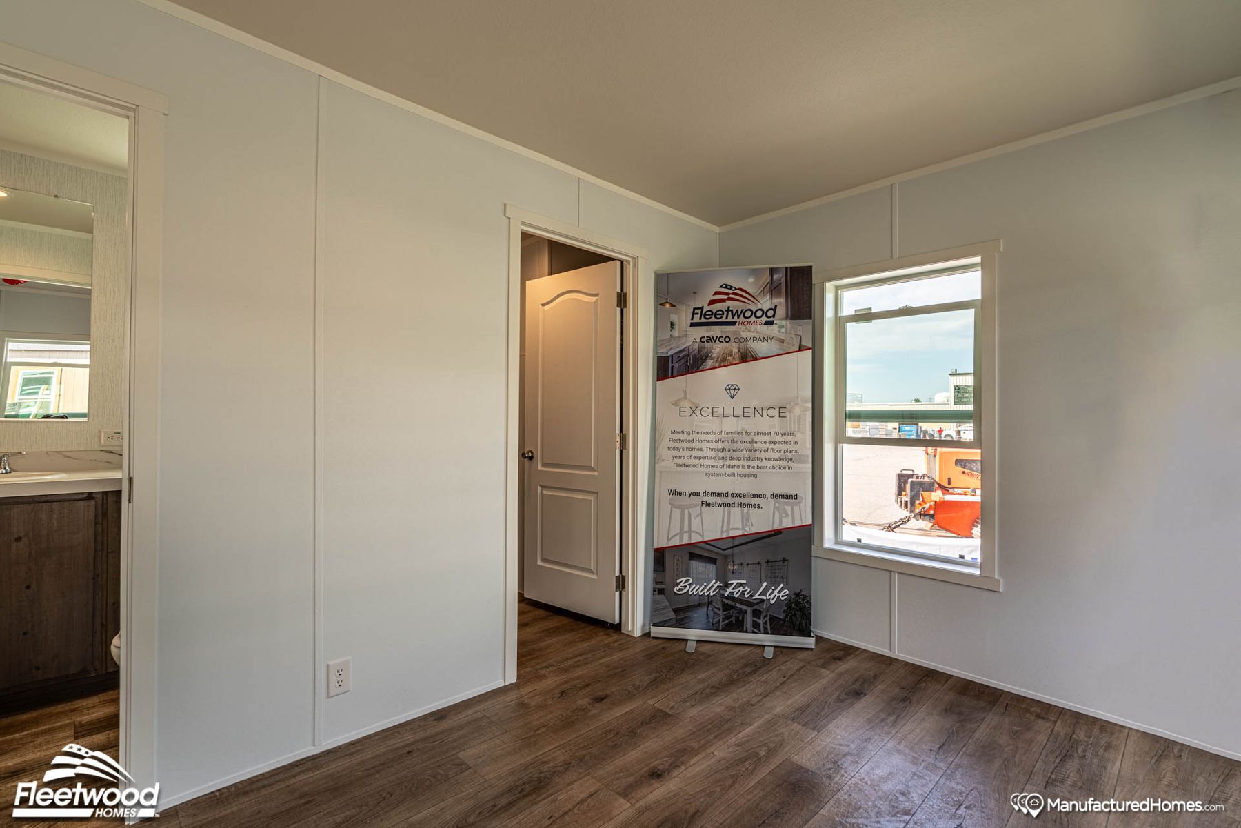 Interior corner of a manufactured home showcasing a Cavco Idaho promotional banner near a window, with wood-look flooring, white walls, and a view into the bathroom and hallway.