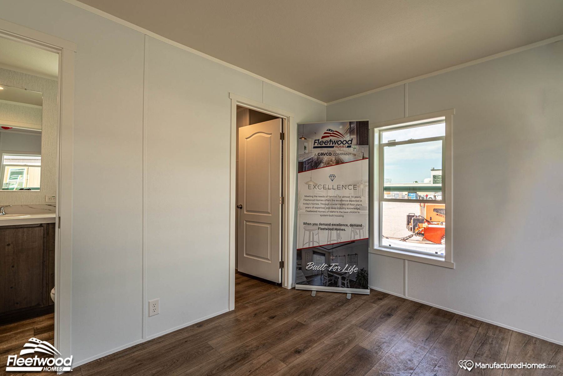 Interior corner of a manufactured home showcasing a Cavco Idaho promotional banner near a window, with wood-look flooring, white walls, and a view into the bathroom and hallway.