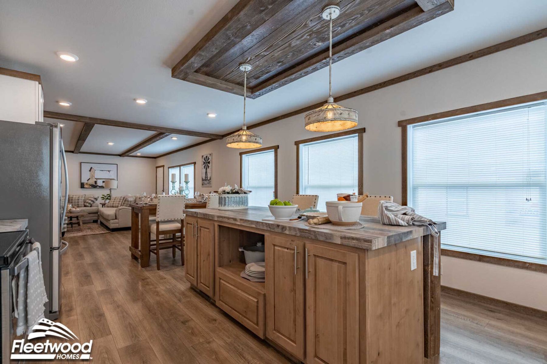 Open-concept kitchen and dining area with a rustic wood island, pendant lighting, wood-look flooring, and large windows, flowing into a living room with a sectional sofa.