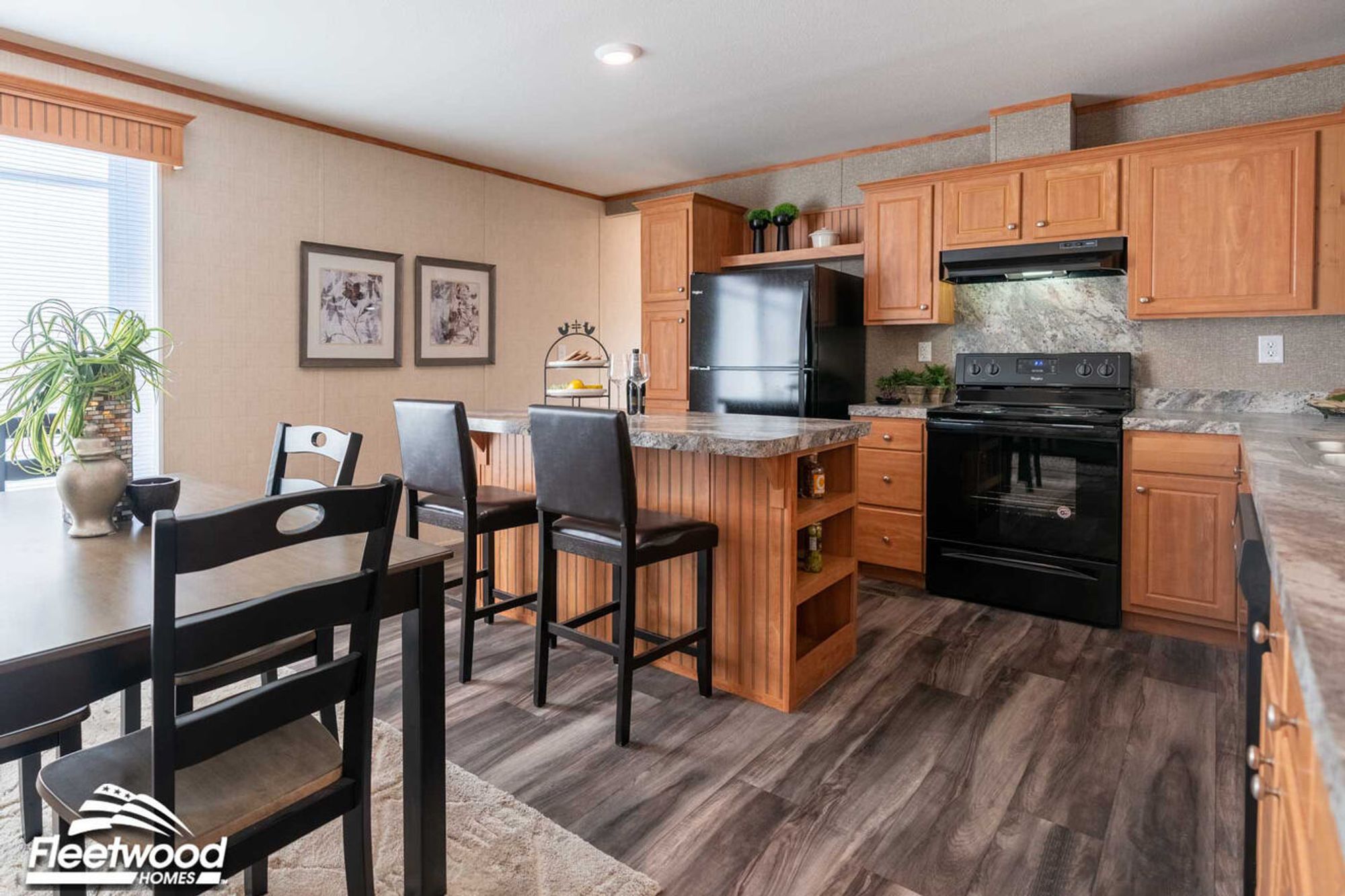 Kitchen with wood cabinetry, a large marble-look island with seating, black appliances including a refrigerator, stove, and dishwasher, and a window above the sink.