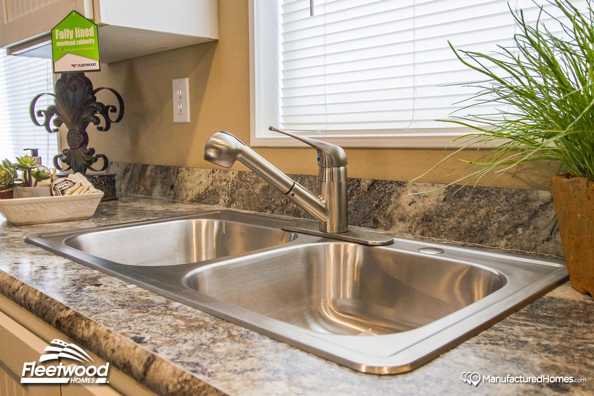 Close-up of a double-basin stainless steel kitchen sink with a pull-out faucet, set in a marbled countertop beneath a window with white blinds.
