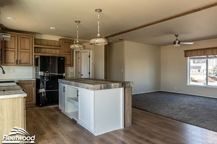 Modern kitchen with wood cabinetry, black appliances, and a rustic island with pendant lighting, opening into a carpeted living area.