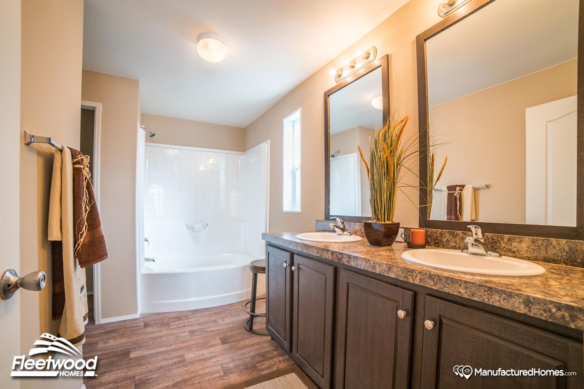 Modern bathroom in a manufactured home featuring a dual-sink vanity with dark wood cabinets, large mirrors, and a white tub-shower combo with curved design.