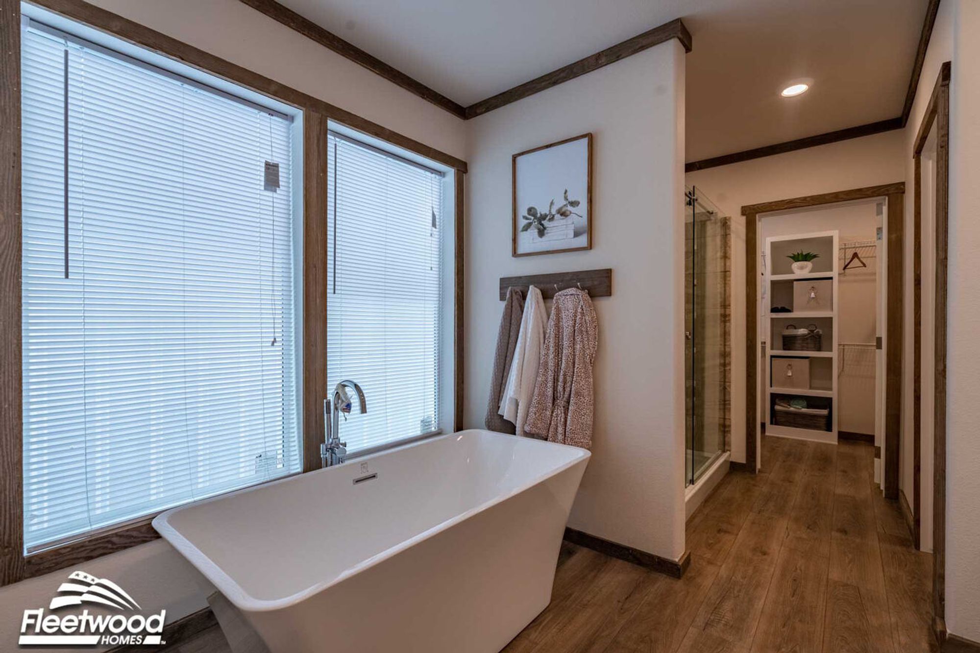 Bathroom with a modern freestanding soaking tub positioned by large windows, wall hooks with robes, and a hallway leading to a walk-in shower and spacious closet with built-in shelving.