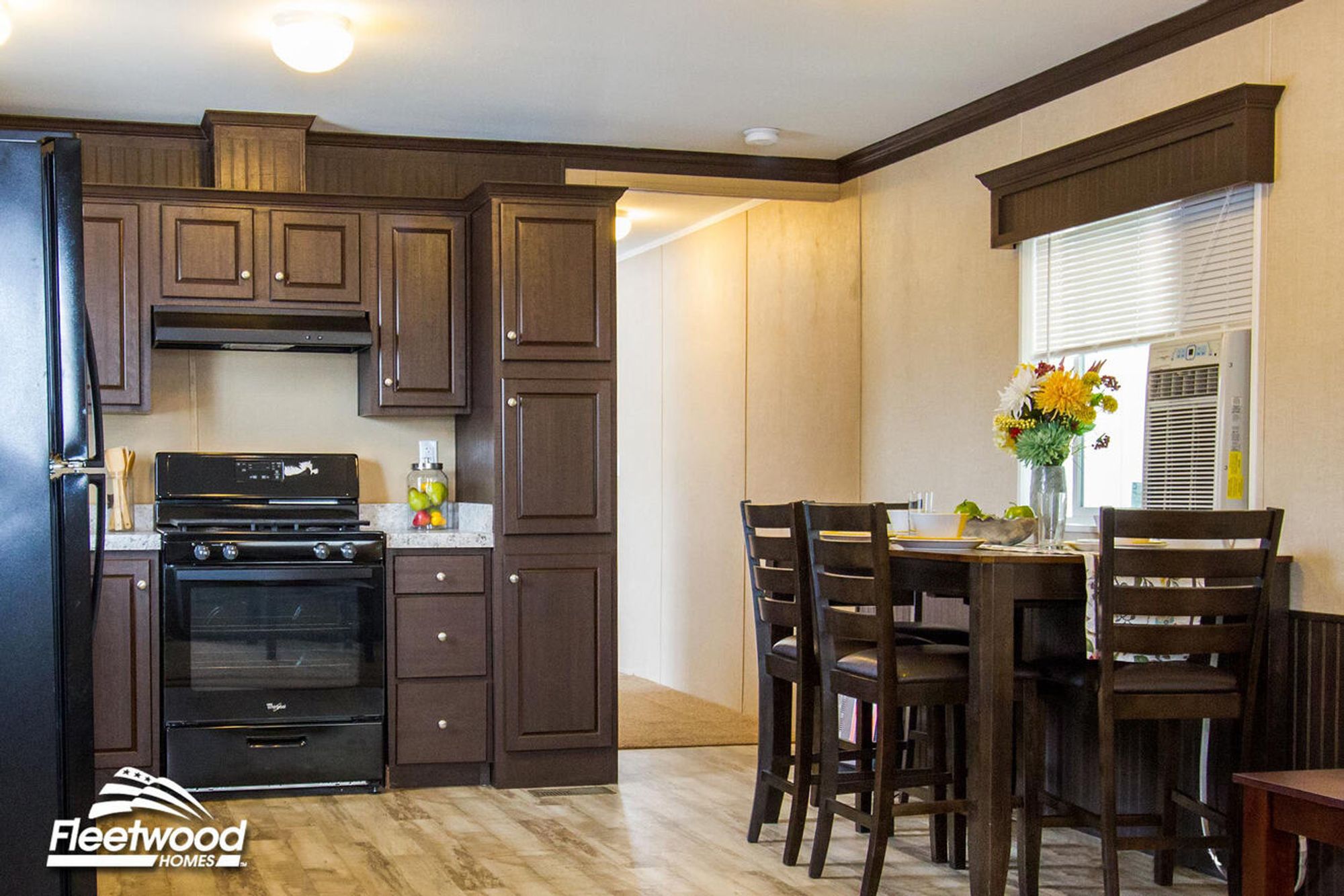 Kitchen with dark brown cabinets, a black stove, and refrigerator, next to a small dining area with a dark wooden table, four chairs, and a vase of flowers by a window with an air conditioning unit.