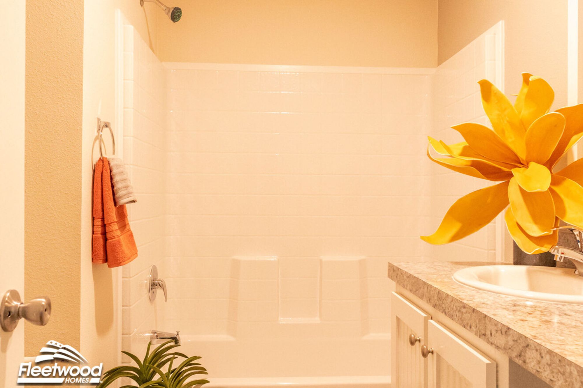 Bright bathroom with a white tiled shower-tub combo, orange towel set, large faux yellow flower decor, and a granite-style vanity countertop.