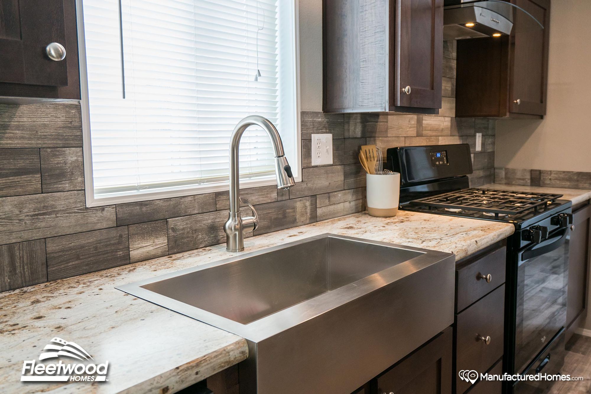 Close-up of a modern kitchen with a stainless steel farmhouse sink, gooseneck faucet, granite countertops, and dark cabinetry with wood-look tile backsplash.
