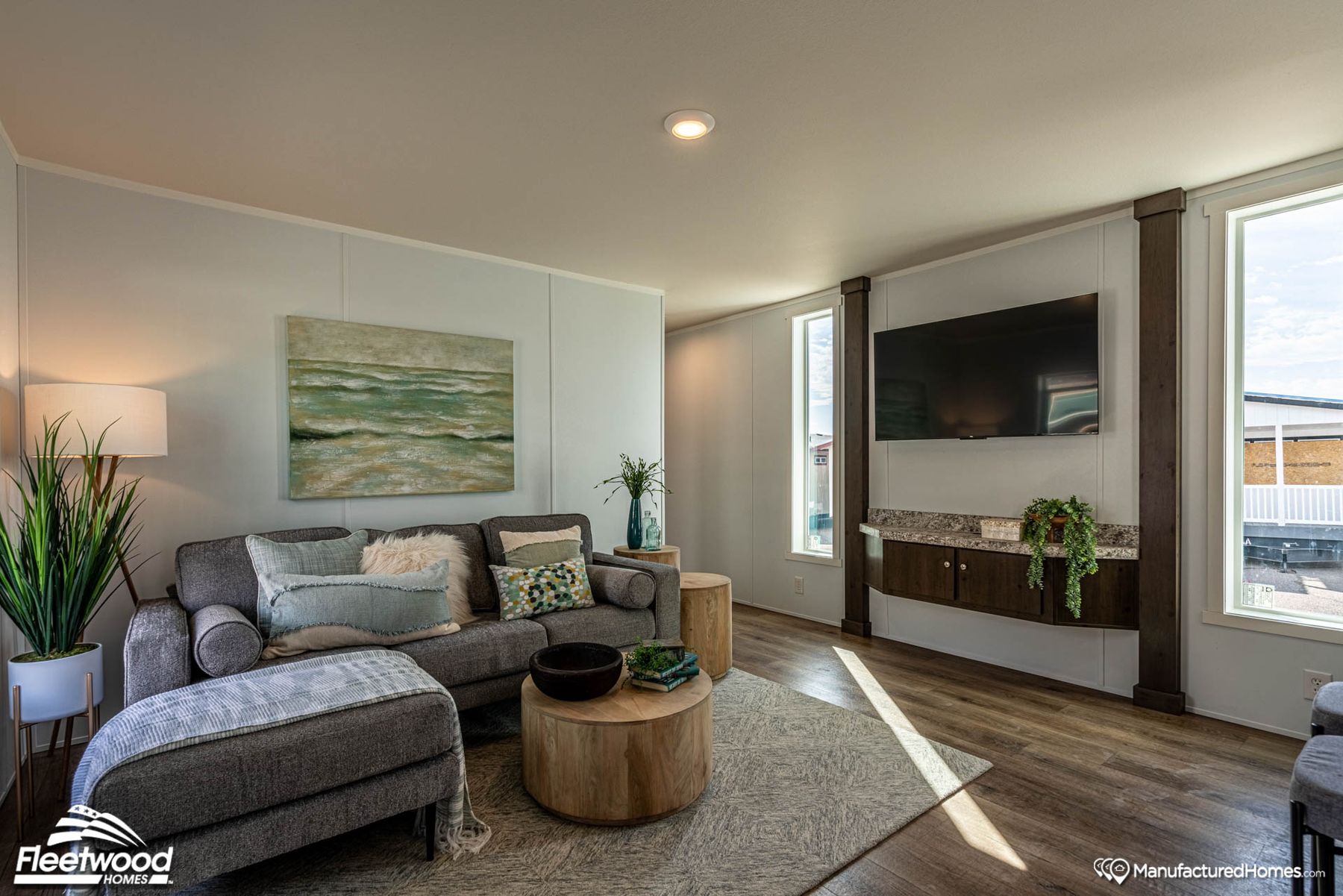 Bright and modern living room with a gray sectional sofa, decorative pillows, round wood coffee tables, and soft natural light from two windows, styled with coastal-themed artwork and plants.