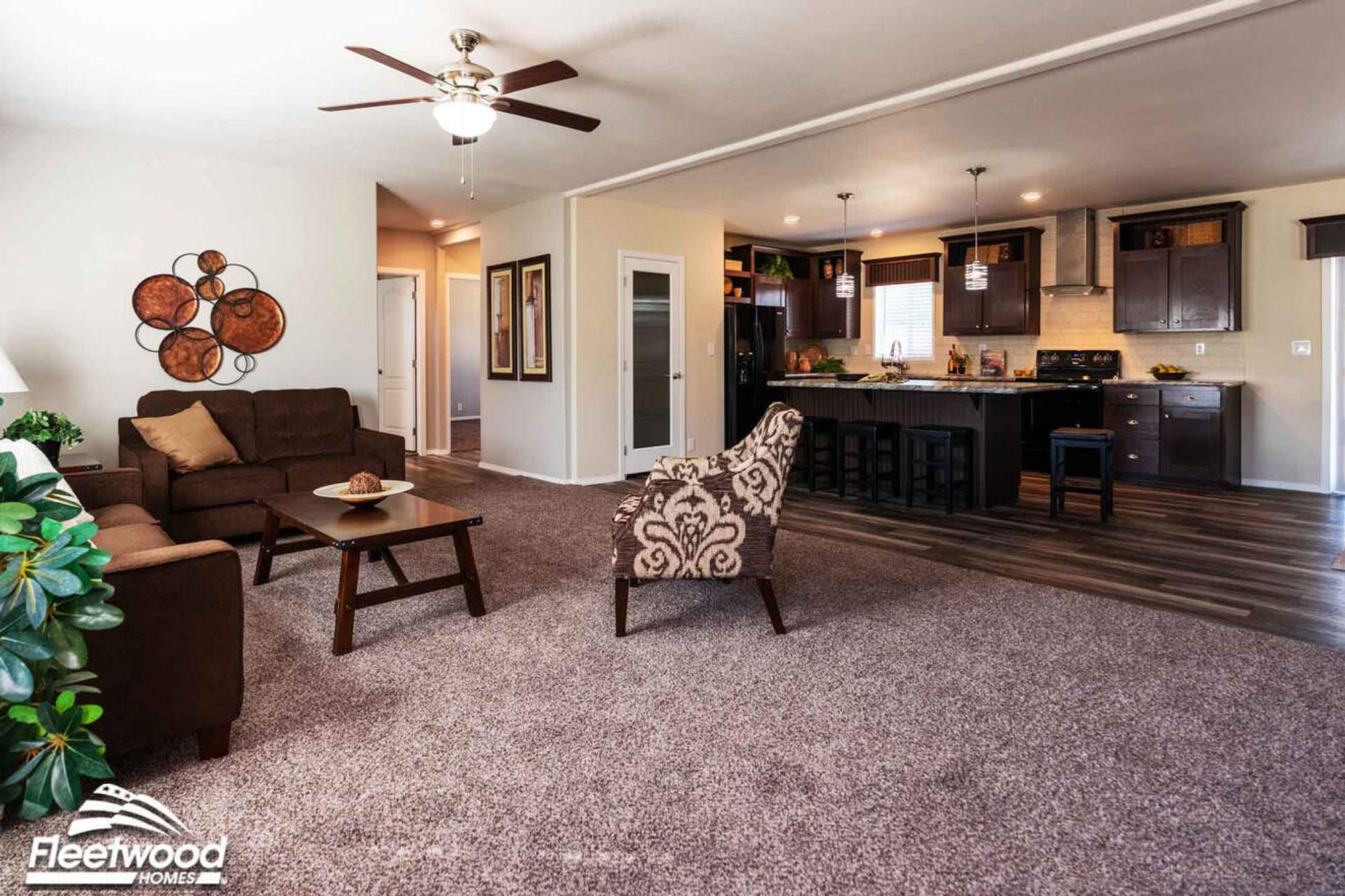 Open living room with brown sofas, patterned accent chair, and view of a modern kitchen with island seating and dark cabinetry.