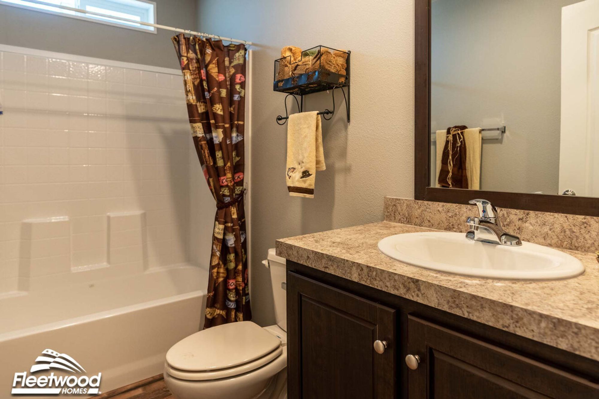 Compact bathroom with a brown patterned shower curtain, beige towels, single-sink vanity with marble countertop, and a wall-mounted wire basket for storage.