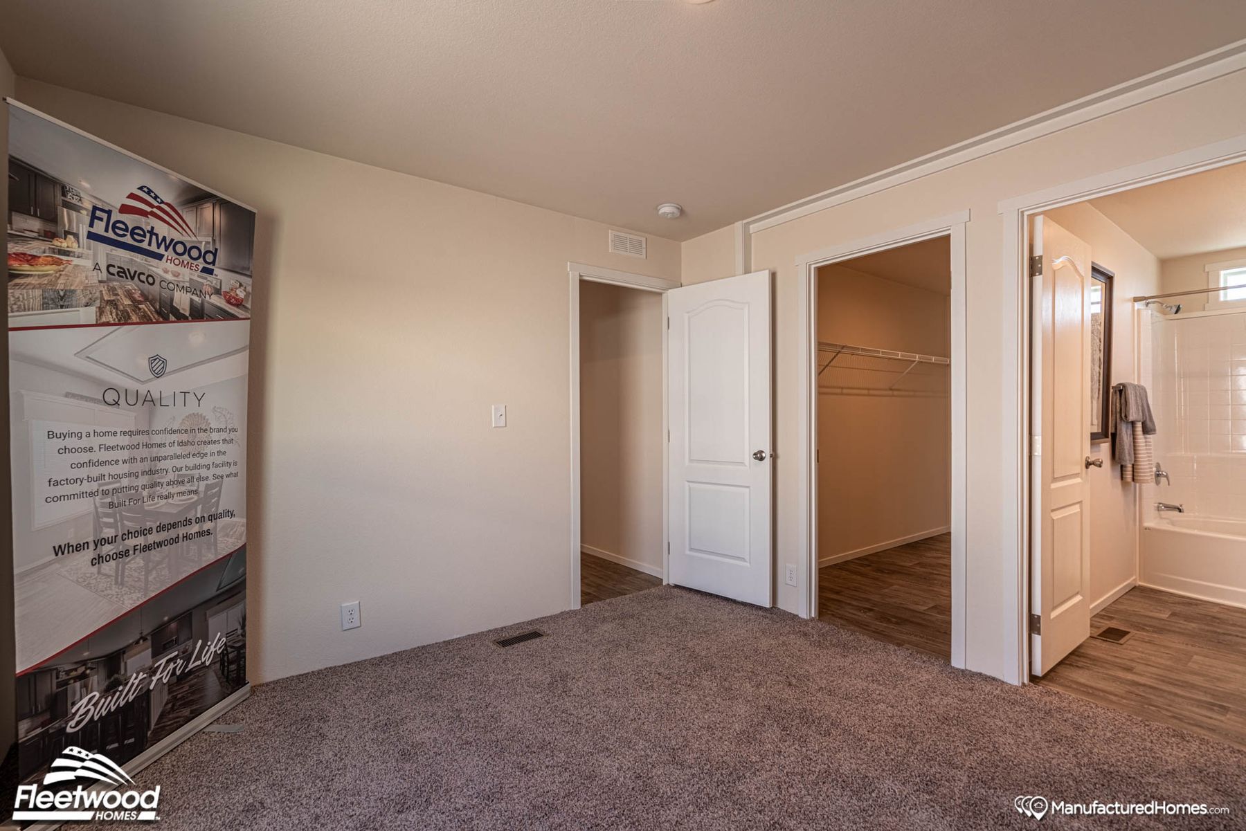 Carpeted bedroom with beige walls, featuring a Cavco Idaho banner, open doors to a walk-in closet and bathroom, and wood-style flooring in adjoining spaces.