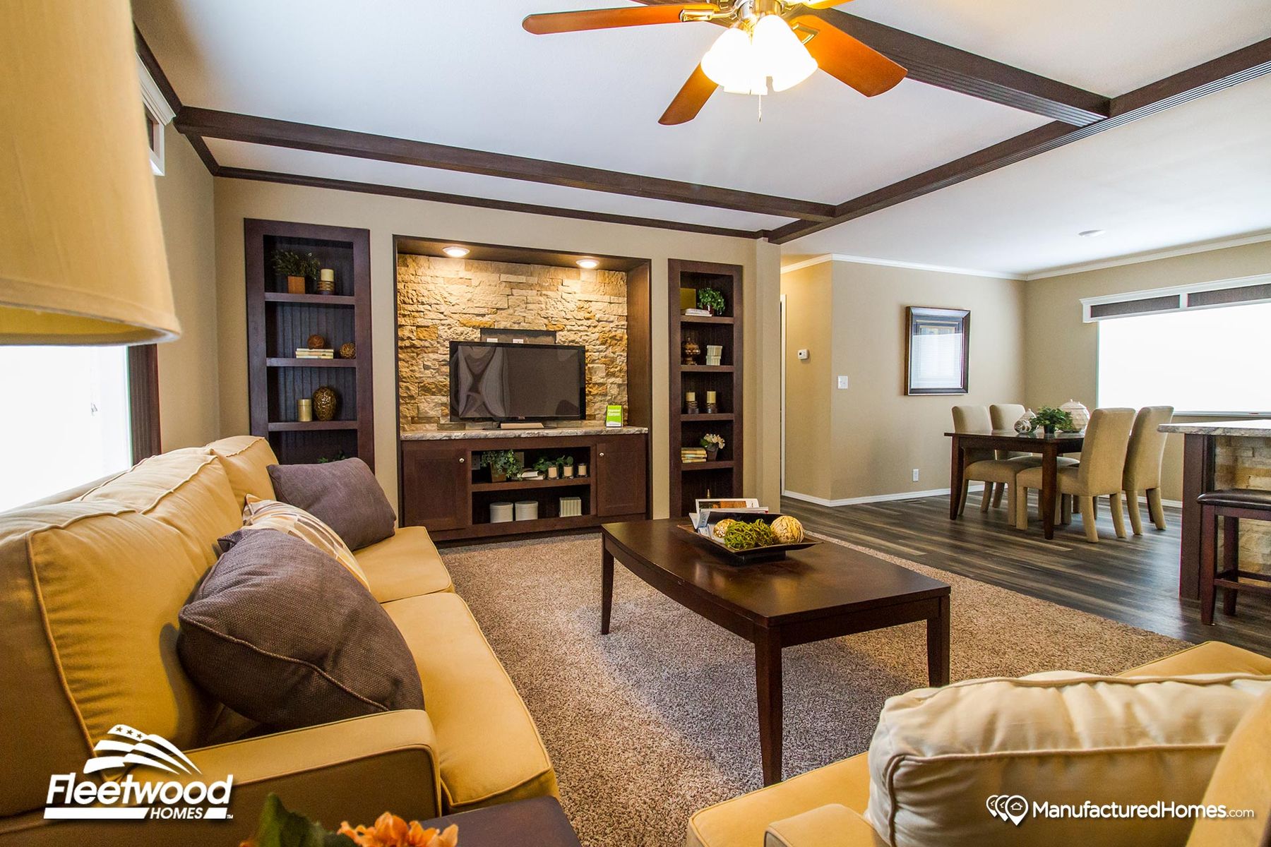 Bright living room in a manufactured home featuring tan sofas with decorative pillows, a dark wood coffee table, carpeted floors, and a ceiling fan with light fixture.