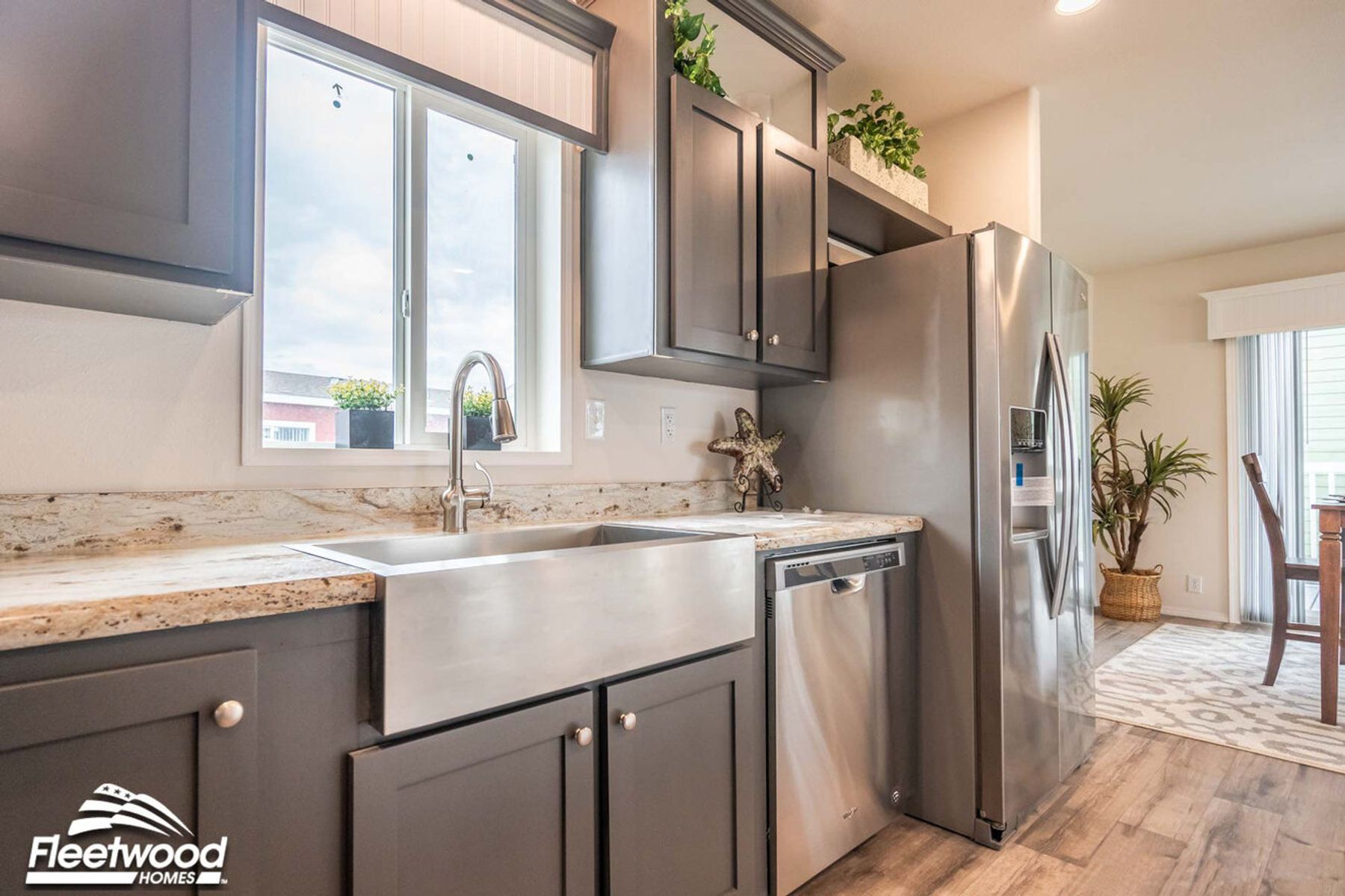 Kitchen with gray cabinets, granite countertops, a stainless steel farmhouse sink, dishwasher, and refrigerator, accented by plants and natural light from a large window.