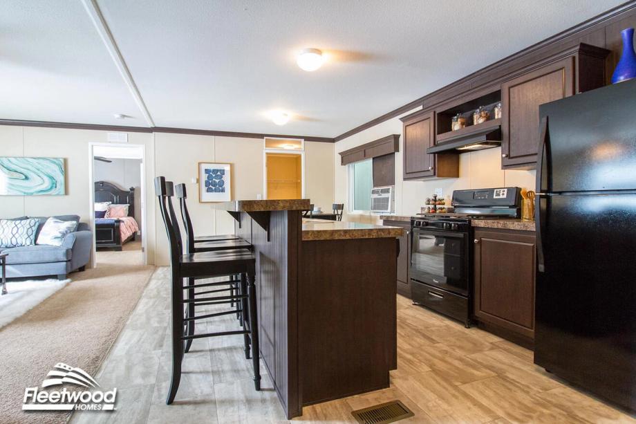 Kitchen with dark wood cabinets, black appliances, and a center island with bar seating, opening into a living area with a sofa and bedroom visible in the background.