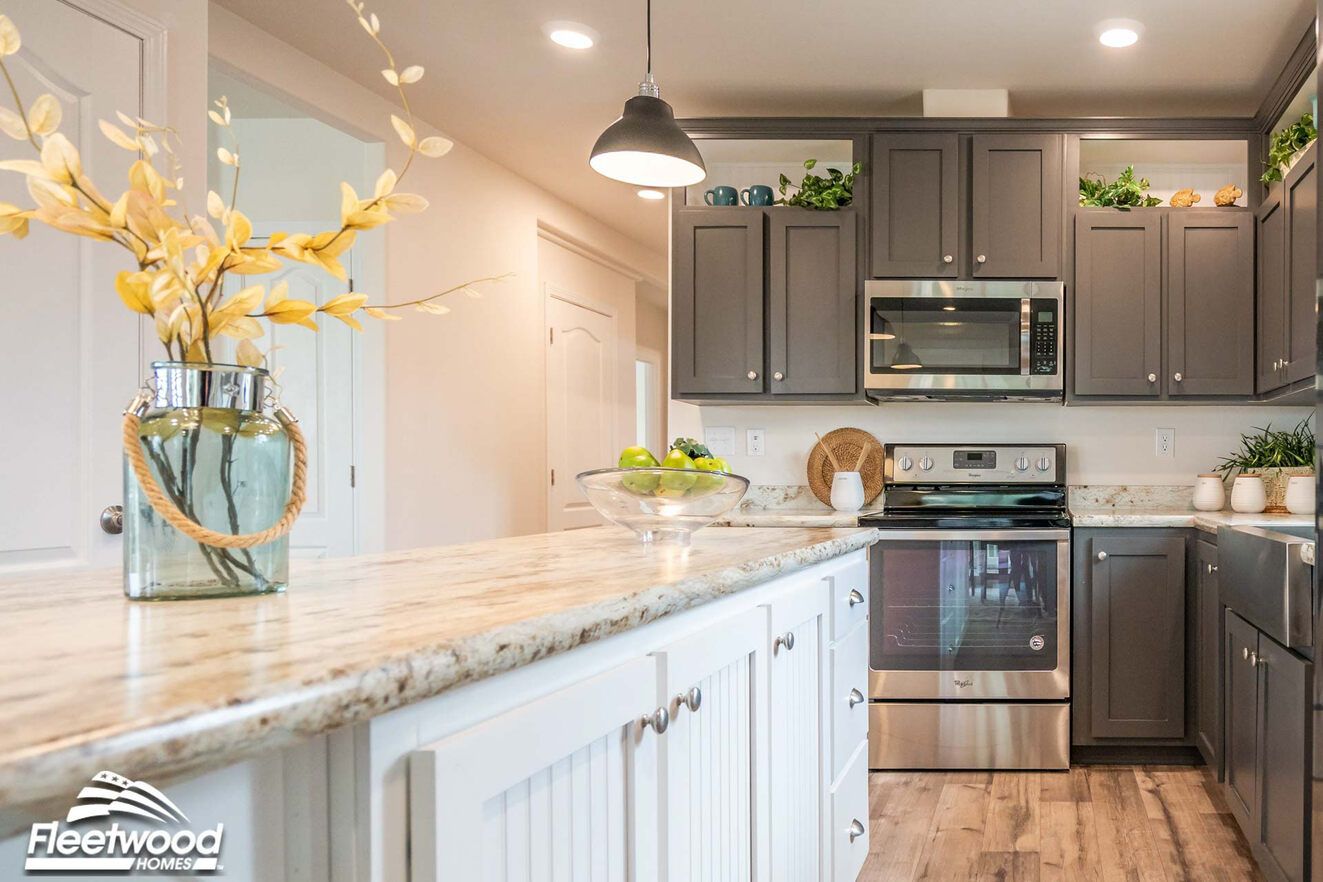 Modern kitchen featuring gray cabinets with stainless steel appliances, including an oven and microwave, granite countertops, and a center island with white cabinetry. Decorative plants, bowls, and a vase with yellow foliage add accents.