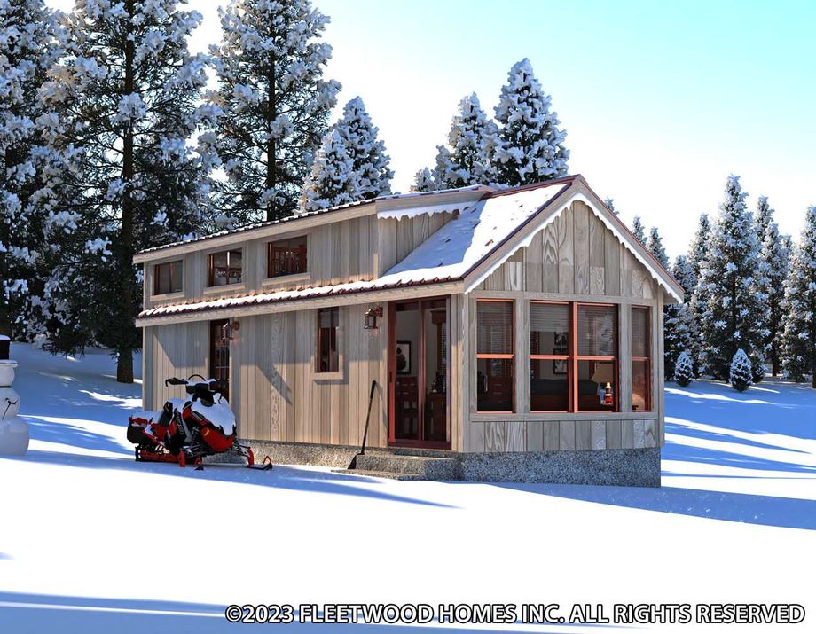 Cabin-style tiny home in a snowy forest setting with wood siding, a pitched roof partially covered in snow, large front windows, and upper loft windows. A snowmobile is parked outside next to a snowman under tall snow-covered pine trees.