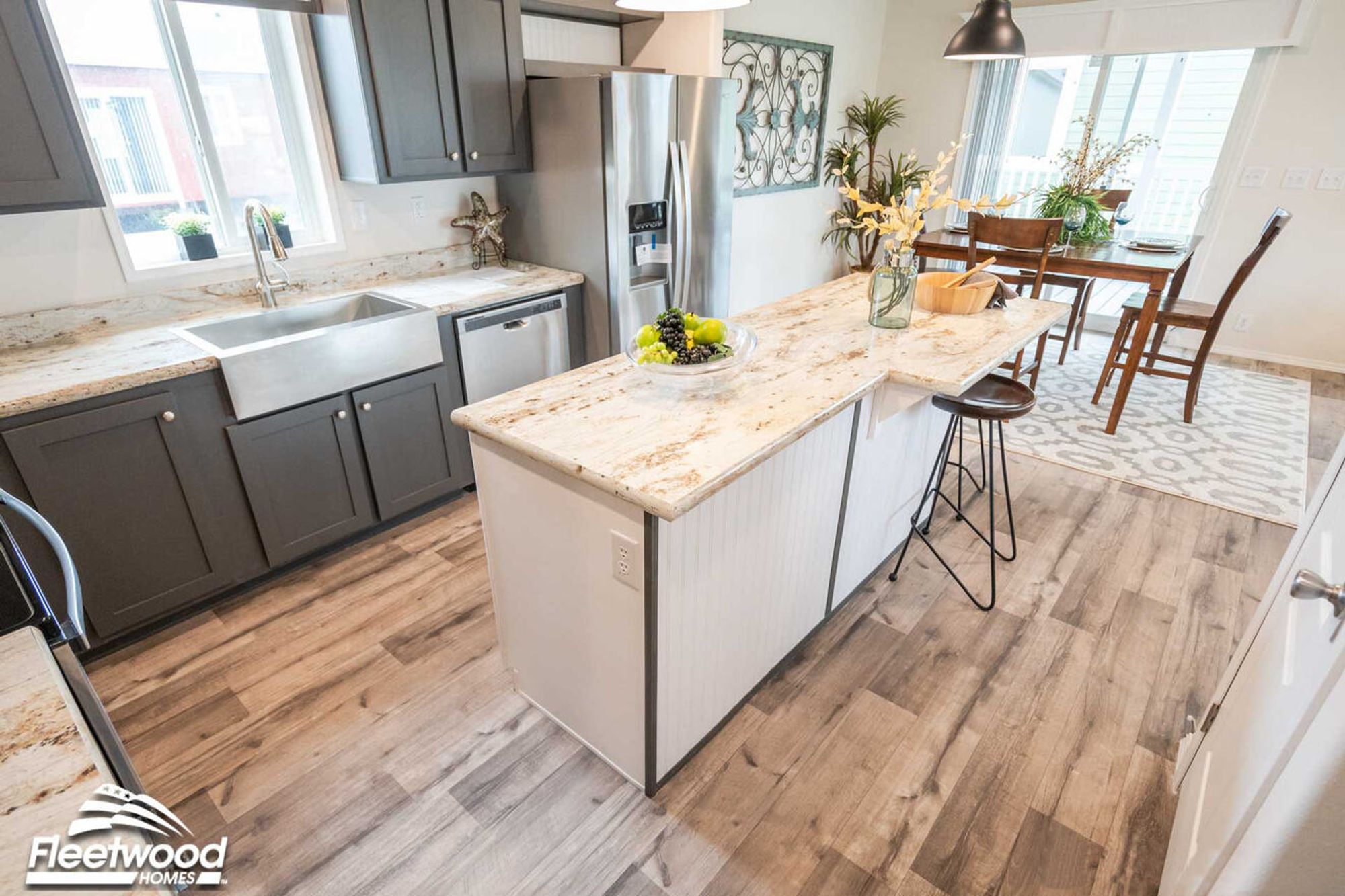 Modern kitchen with gray cabinets, stainless steel farmhouse sink, granite countertops, center island with bar seating, and dining area in view.