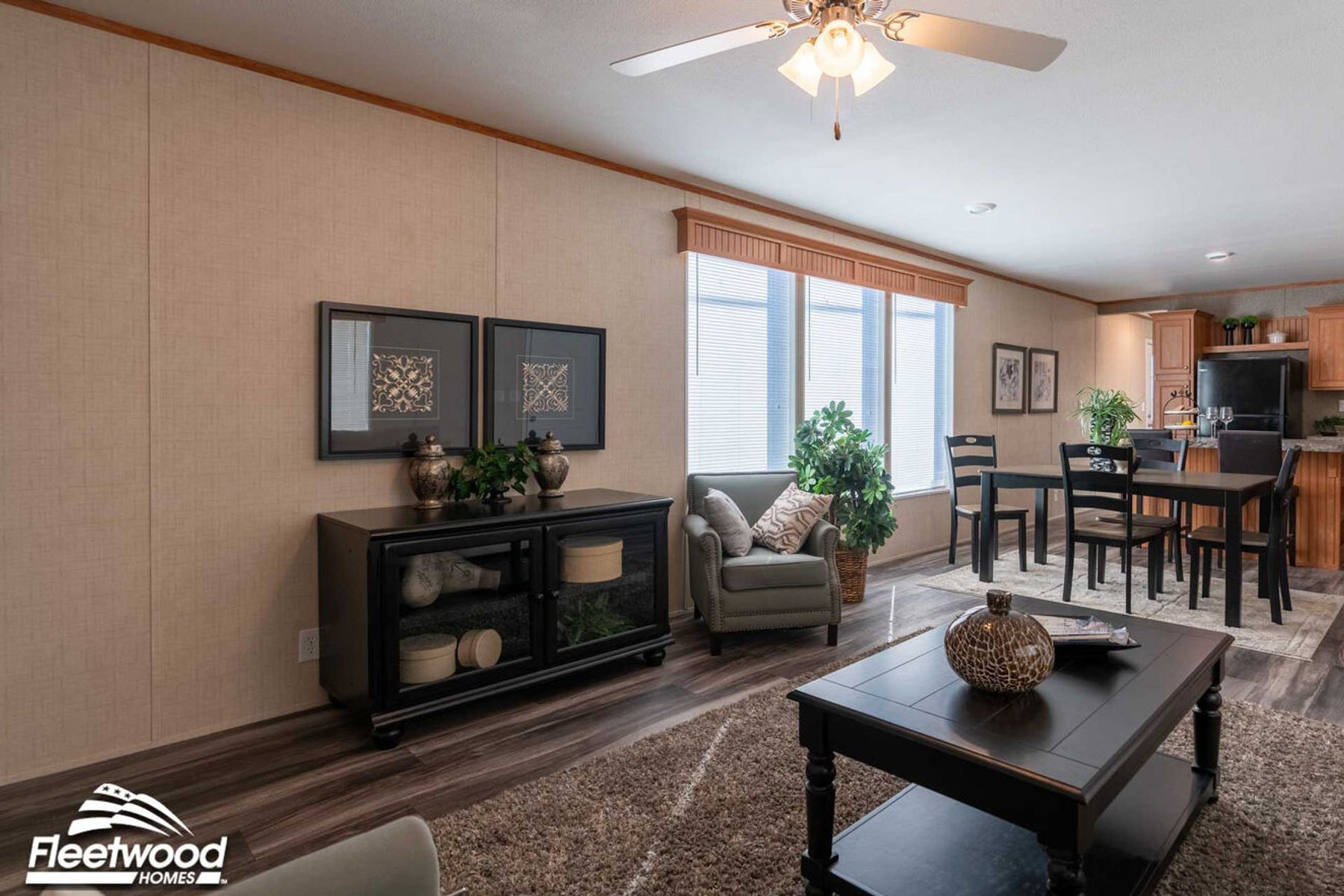 Living and dining area with dark wood furniture, a black cabinet, armchair, and coffee table, decorated with plants and wall art, opening into a kitchen with wood cabinets and black appliances.