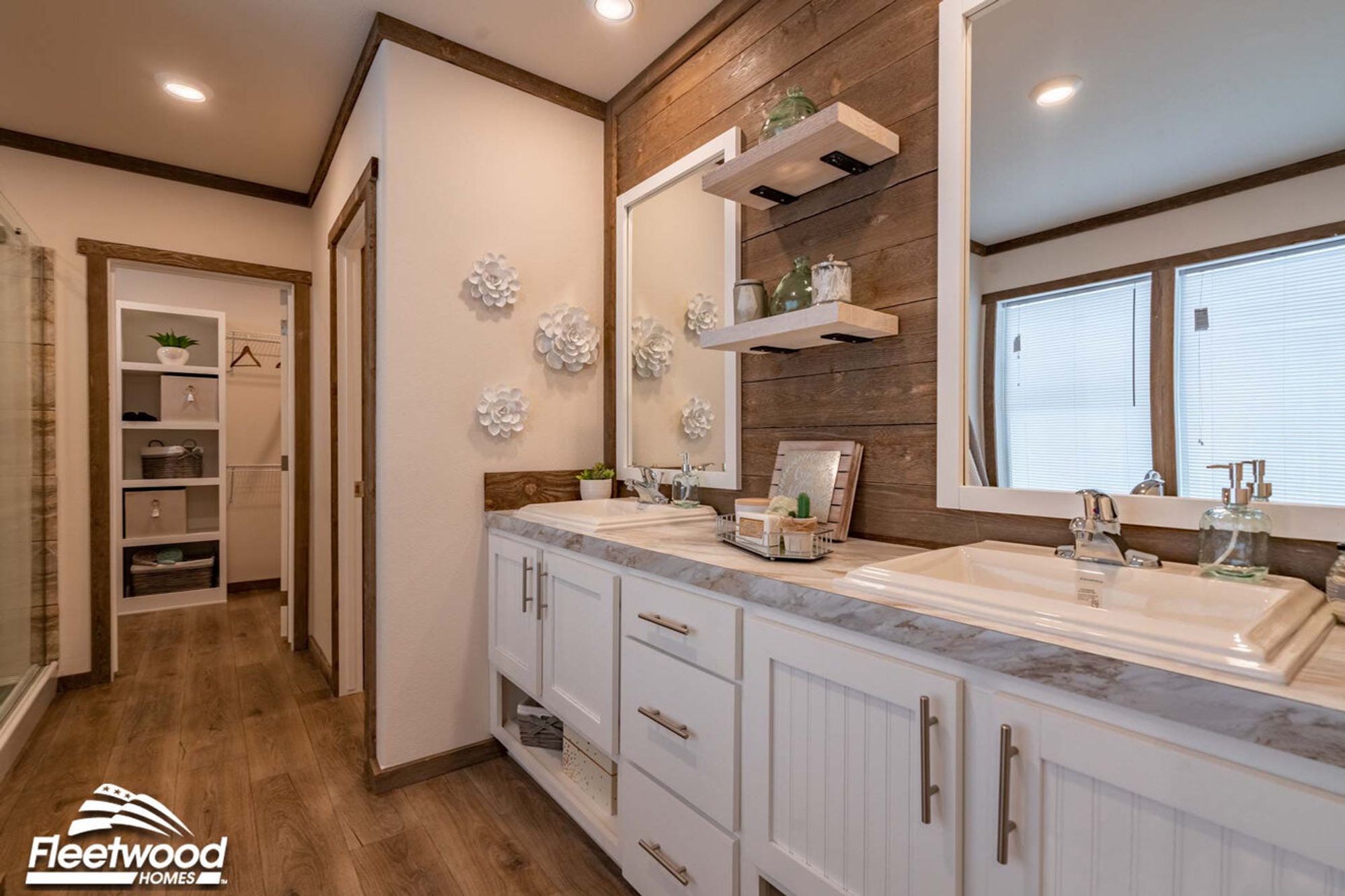 Bathroom vanity with dual sinks, marble-style countertops, white cabinetry, two framed mirrors, wood accent wall, and floating shelves with decorative items.