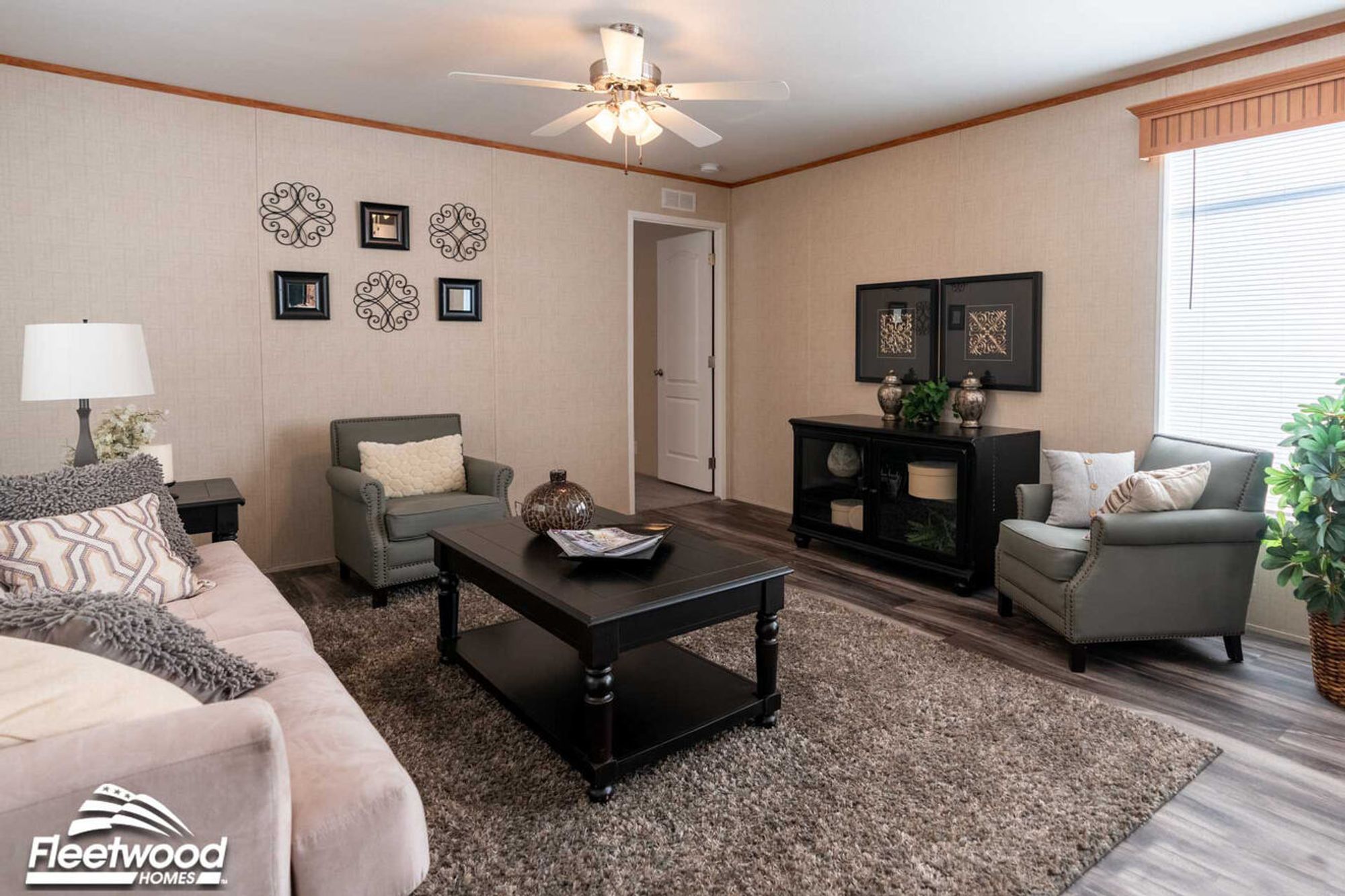 Cozy living room with a beige sofa, two gray armchairs, a dark wood coffee table, and wall art accents under a ceiling fan.